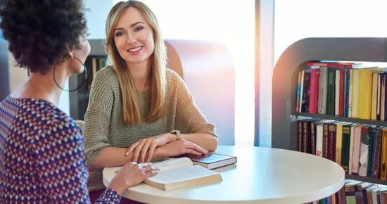 duas mulheres sentadas ao redor de uma mesa conversando sobre as funções de uma corretora de consórcios